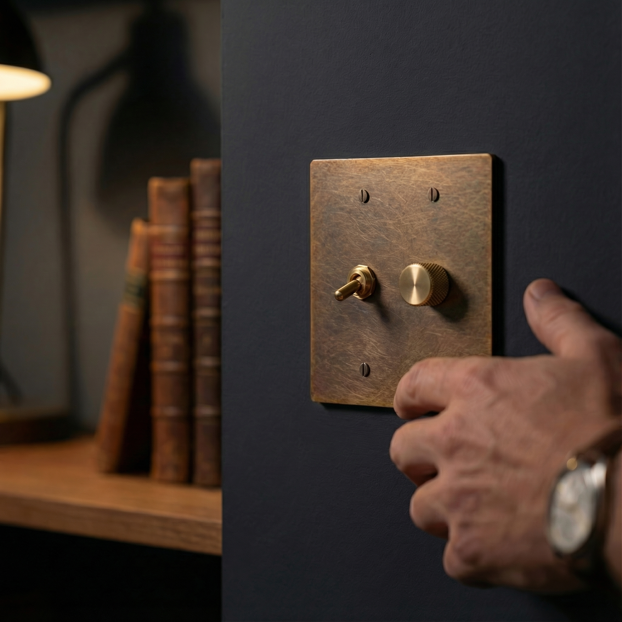 A hand adjusts the aged brass 2 gang dimmer and vintage toggle switch plate in a timeless, sophisticated study.
