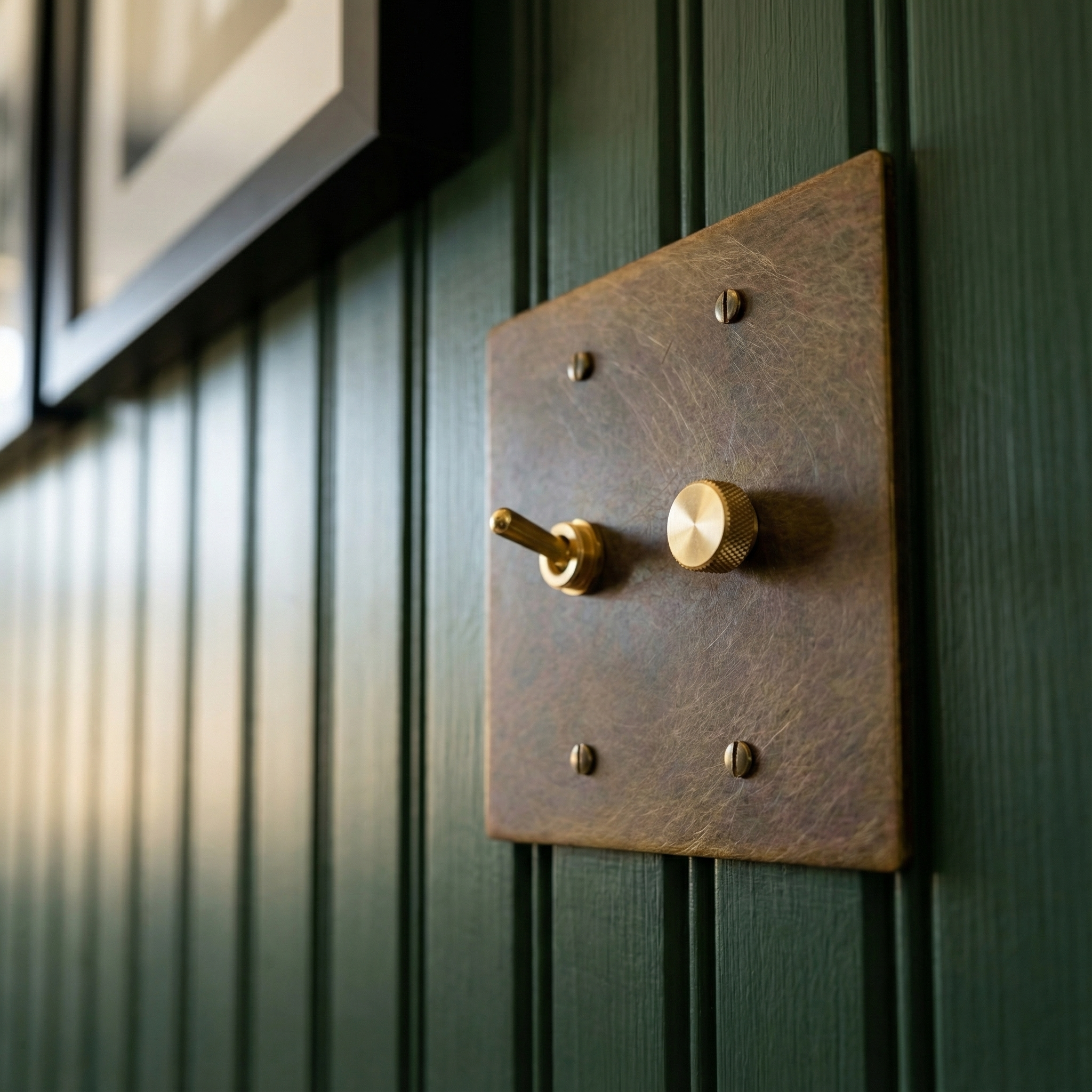 Aged brass 2 gang dimmer and vintage toggle switch plate on a deep green paneled wall, exuding timeless elegance.