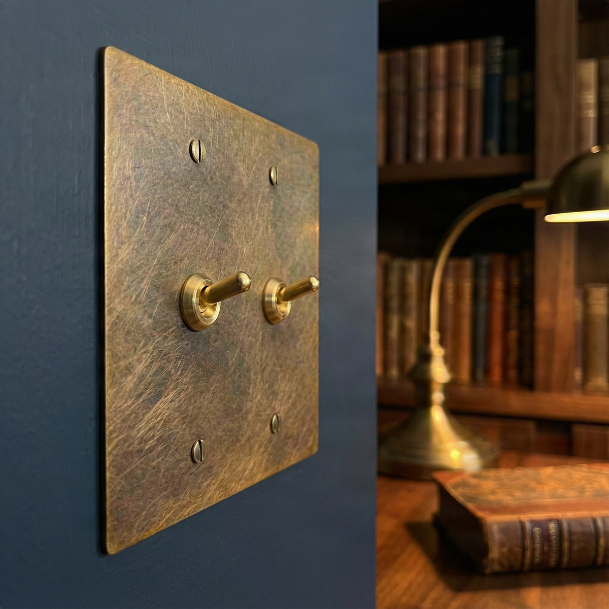 Aged brass 2 gang vintage toggle switch plate on a dark wall, complementing a timeless study with books and a lamp.