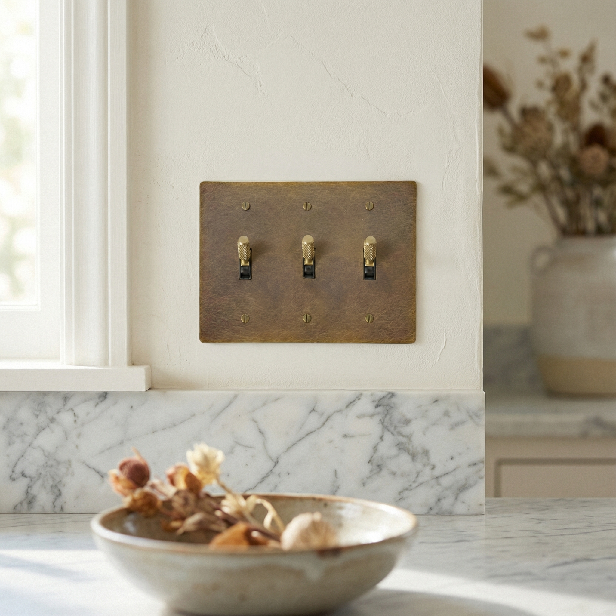 Timeless aged brass 3 gang toggle switch plate with knurled toggles, elegant on a textured wall and marble.