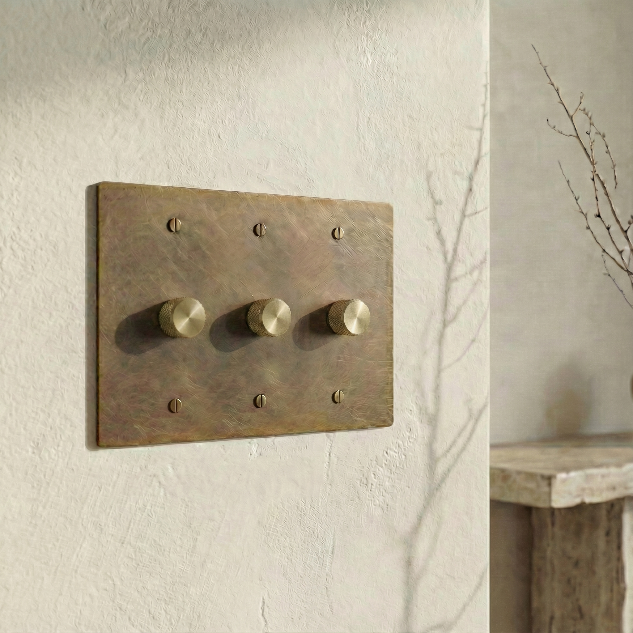 Aged brass 3 gang dimmer switch plate, knurled controls on a textured wall with subtle branch shadows, quiet luxury.