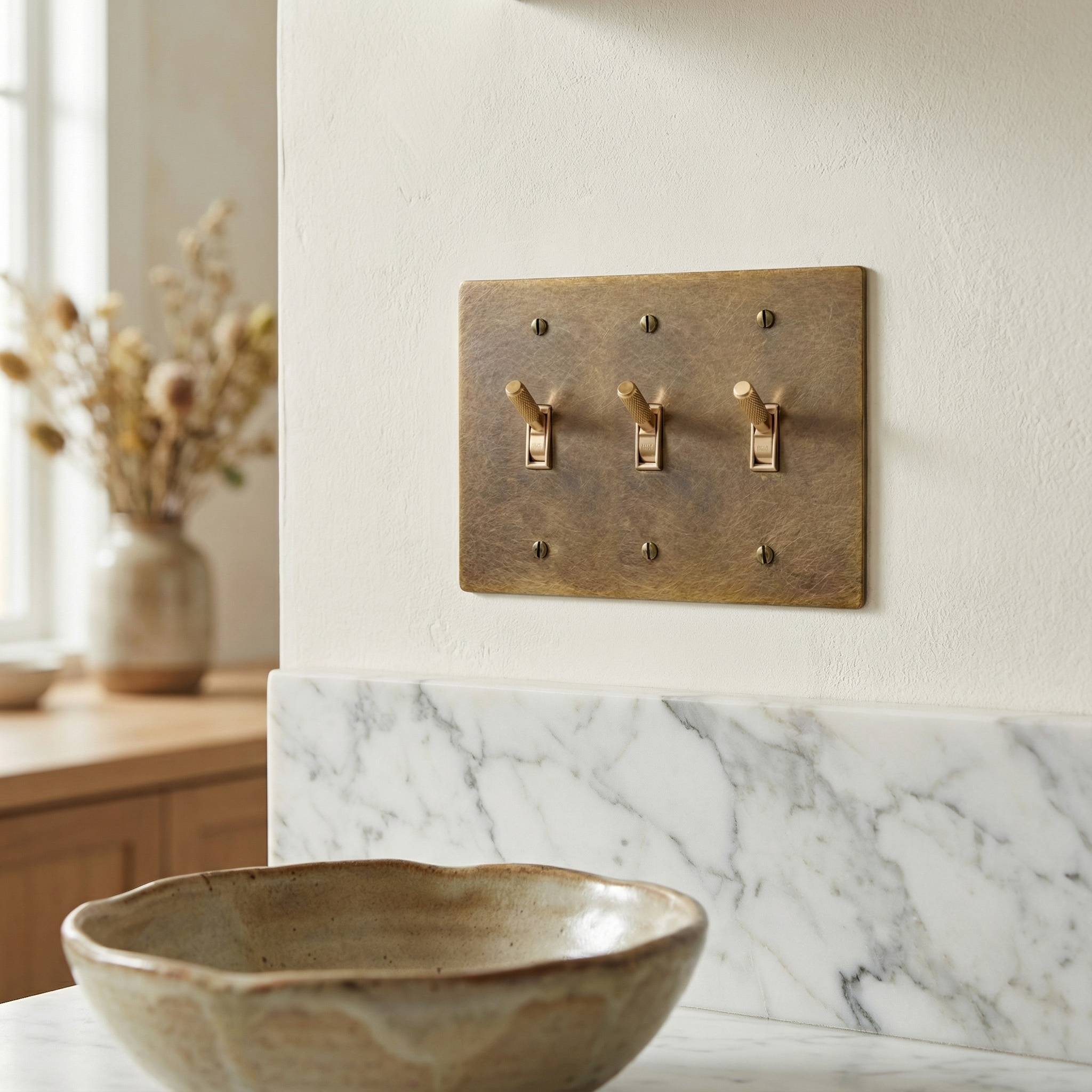 Aged brass 3 gang toggle switch plate on a textured cream wall above a white marble counter, conveying quiet luxury.