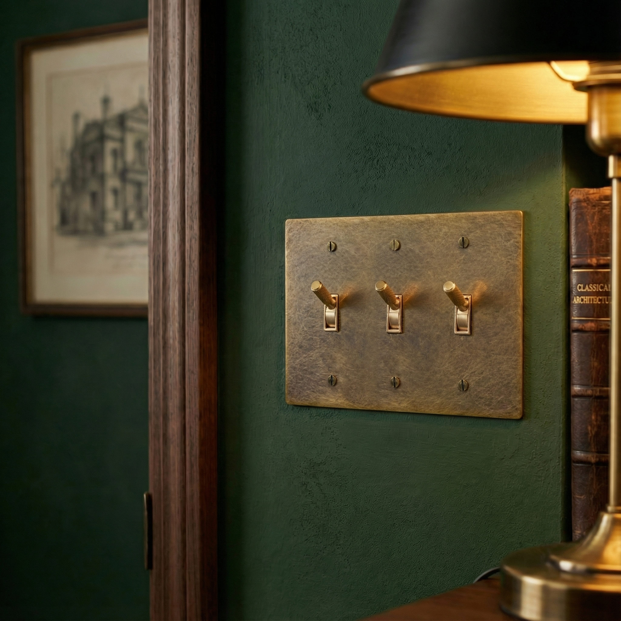 Elegant aged brass 3 gang toggle switch plate on a deep green wall, beside a lamp and classic books.