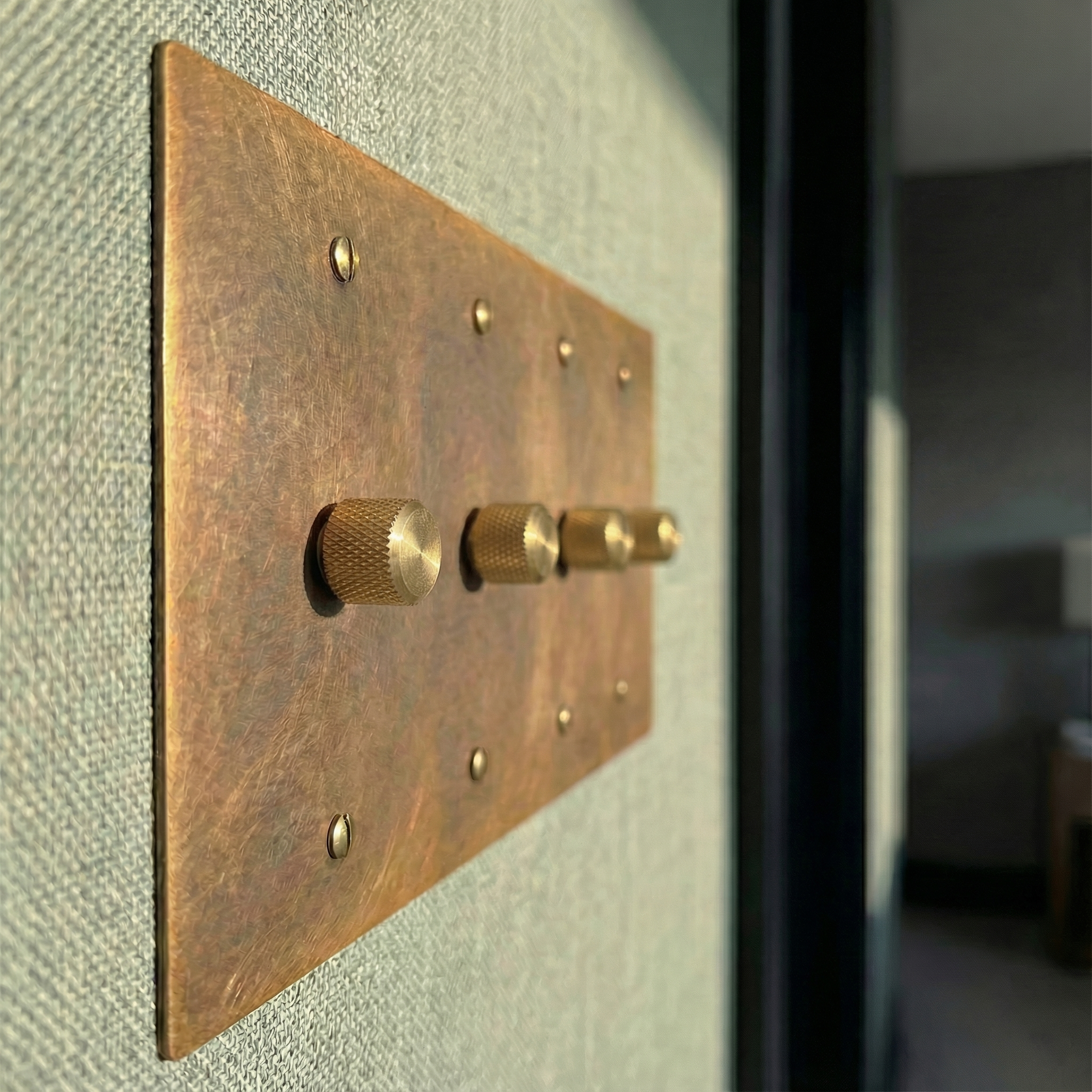 Tactile aged brass 4 gang dimmer switch plate on a woven wall, evoking quiet luxury and enduring craftsmanship.