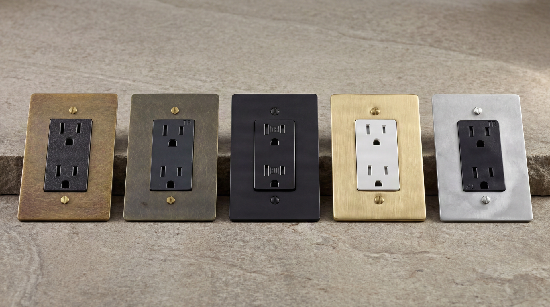 Five authentic Duplex Outlets: aged brass, antique brass, matte black, satin brass, and satin nickel on stone.