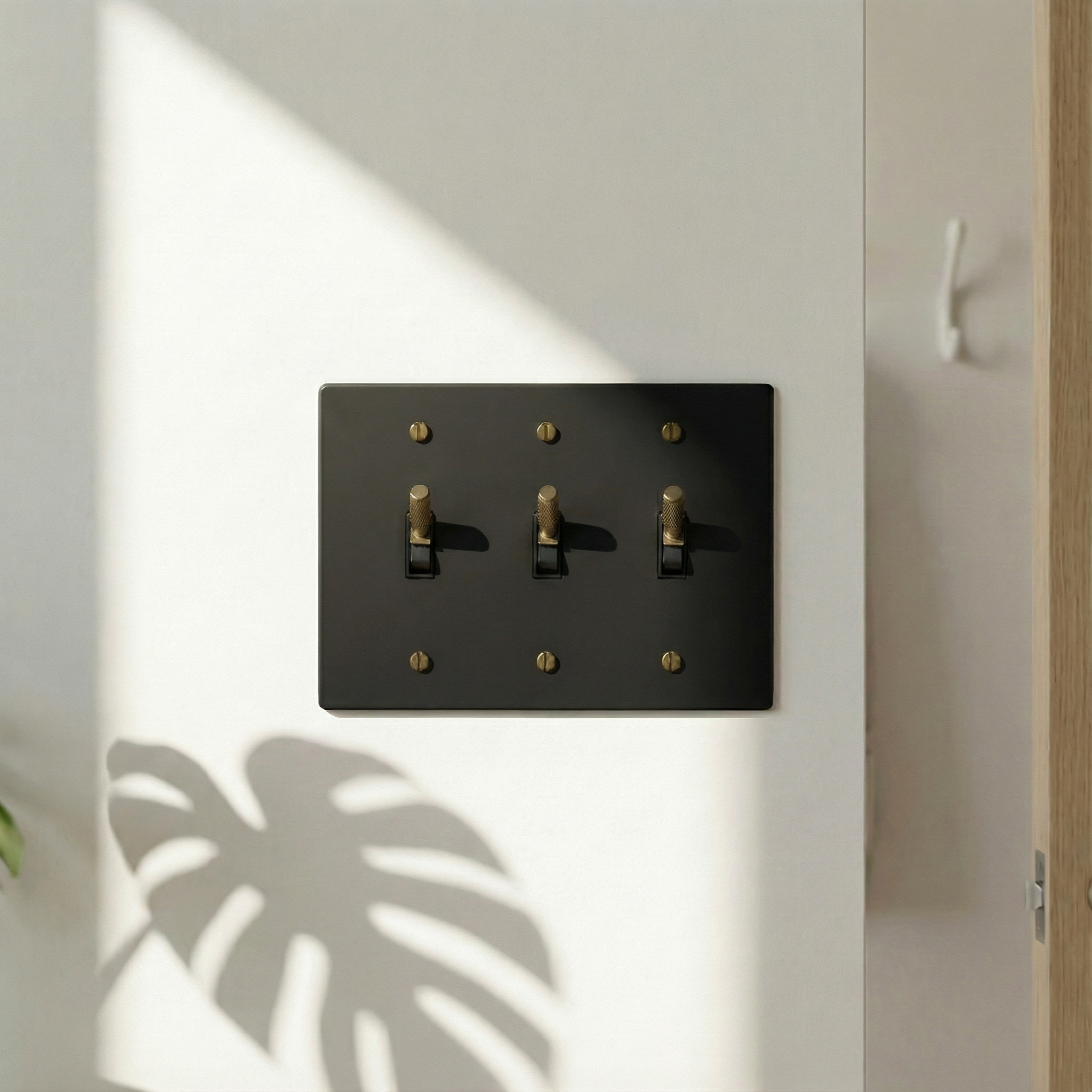 matte black (antique brass accents) 3 gang toggle switch plate exuding quiet luxury on a sunlit wall with a plant shadow.
