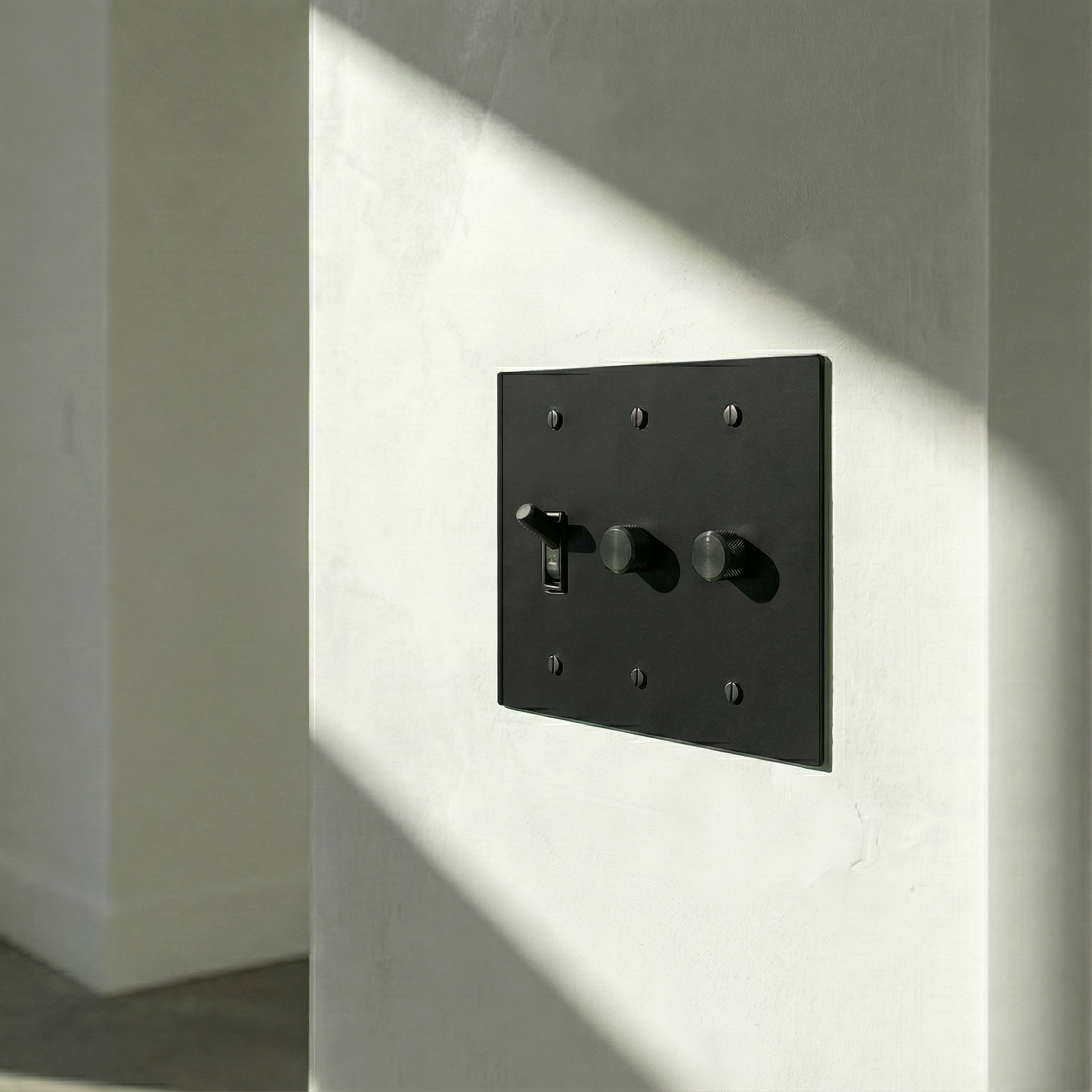 Timeless matte black (matte black accents) 3 gang dimmer and toggle switch plate on a minimalist wall with a sunlight shadow.