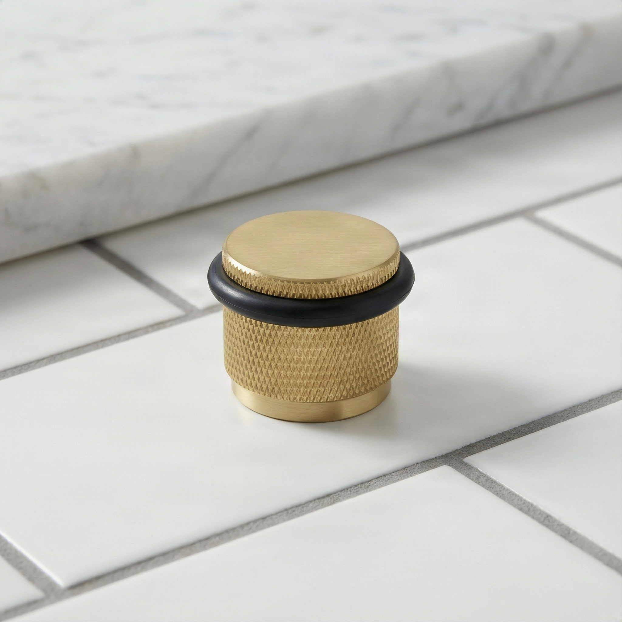 knurled satin brass floor mount door stop on white tile with marble, exuding quiet luxury.