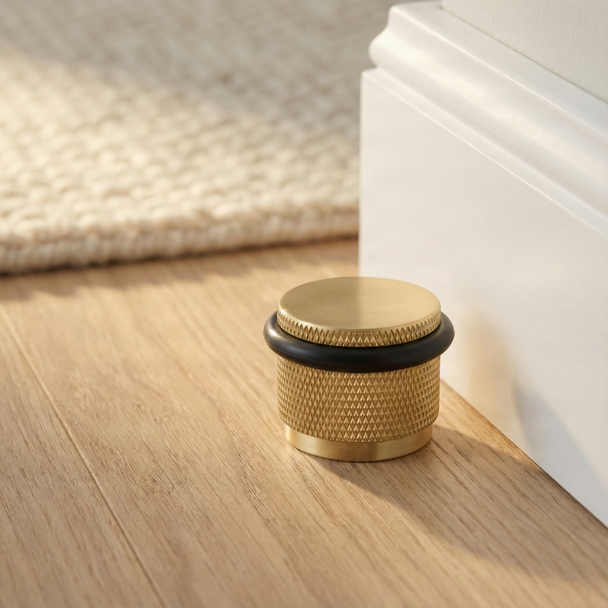 Modern satin brass floor mount door stop, knurled, on wood floor by white trim and a soft rug, evoking quiet luxury.