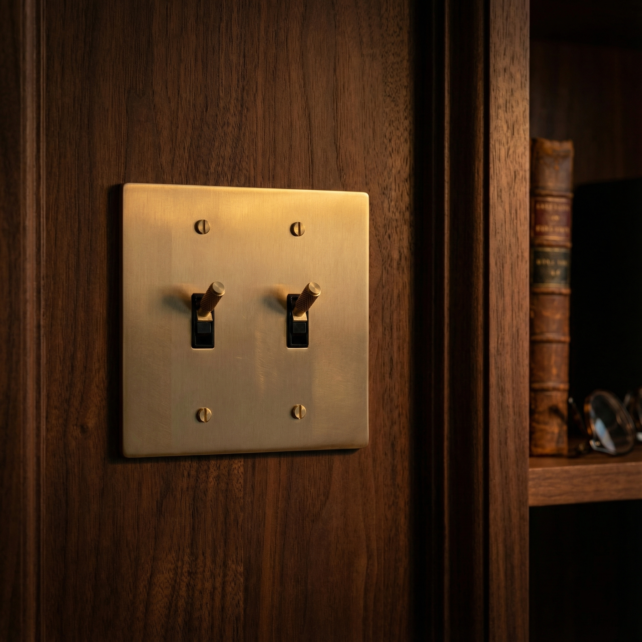 Satin brass 2 gang toggle switch plate on dark wood, next to a bookshelf, evoking timeless elegance.