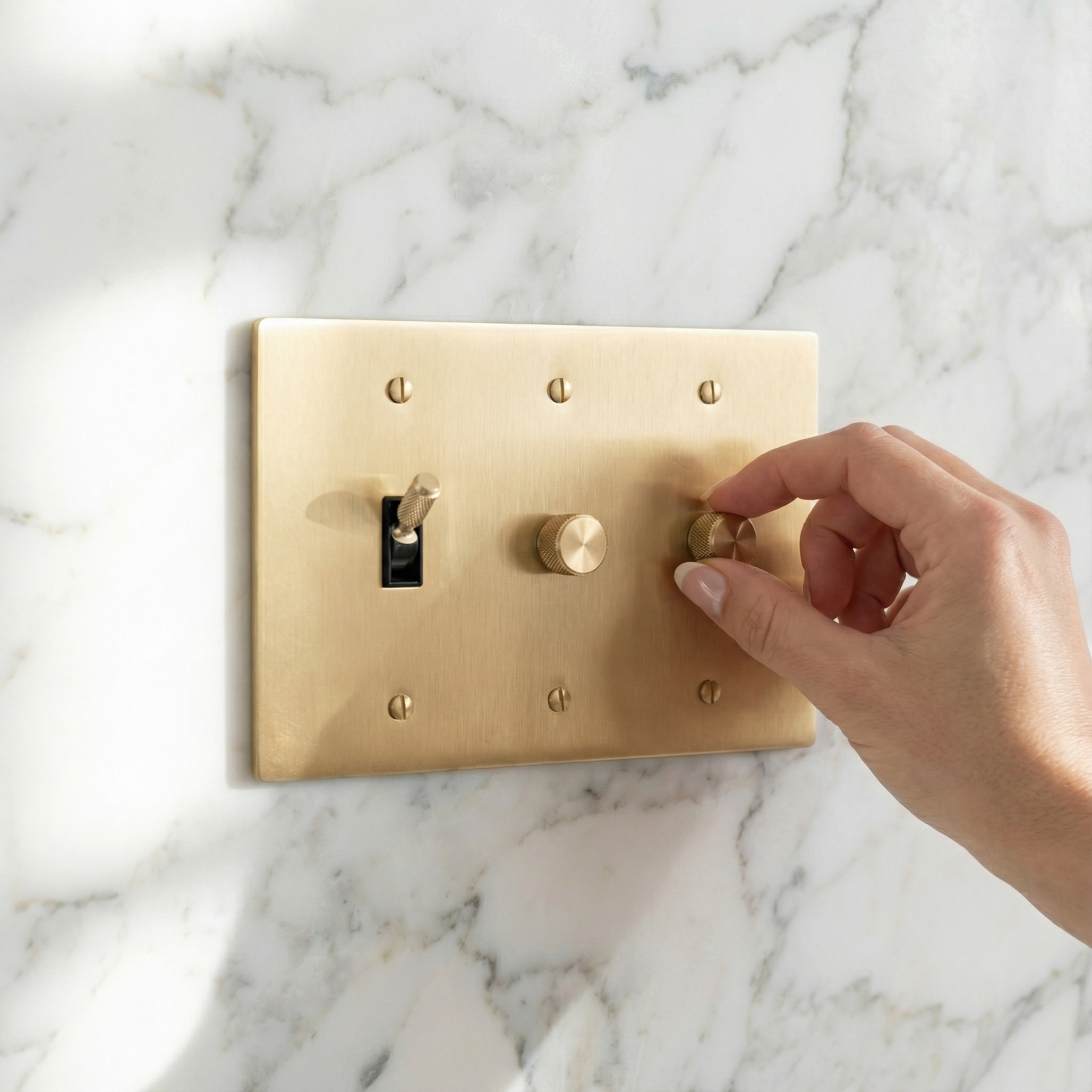 A hand adjusts the satin brass 3 gang dimmer and toggle switch plate on a luxurious marble wall, evoking timeless elegance.