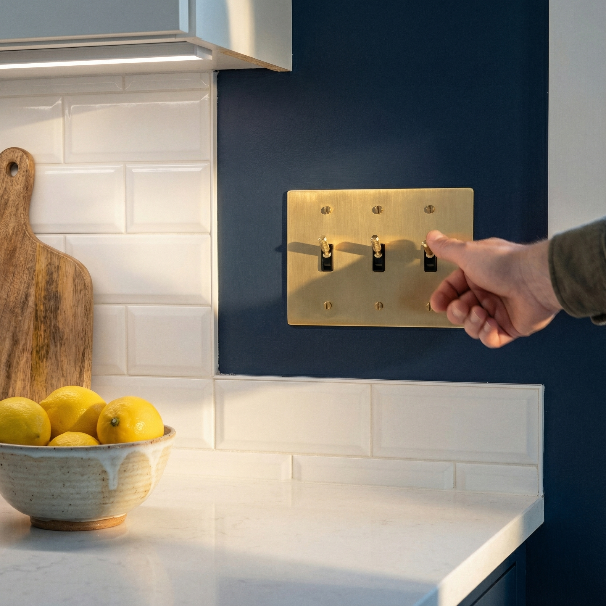 Hand turning a timeless satin brass 3 gang toggle switch plate on a deep blue wall, elegant kitchen.