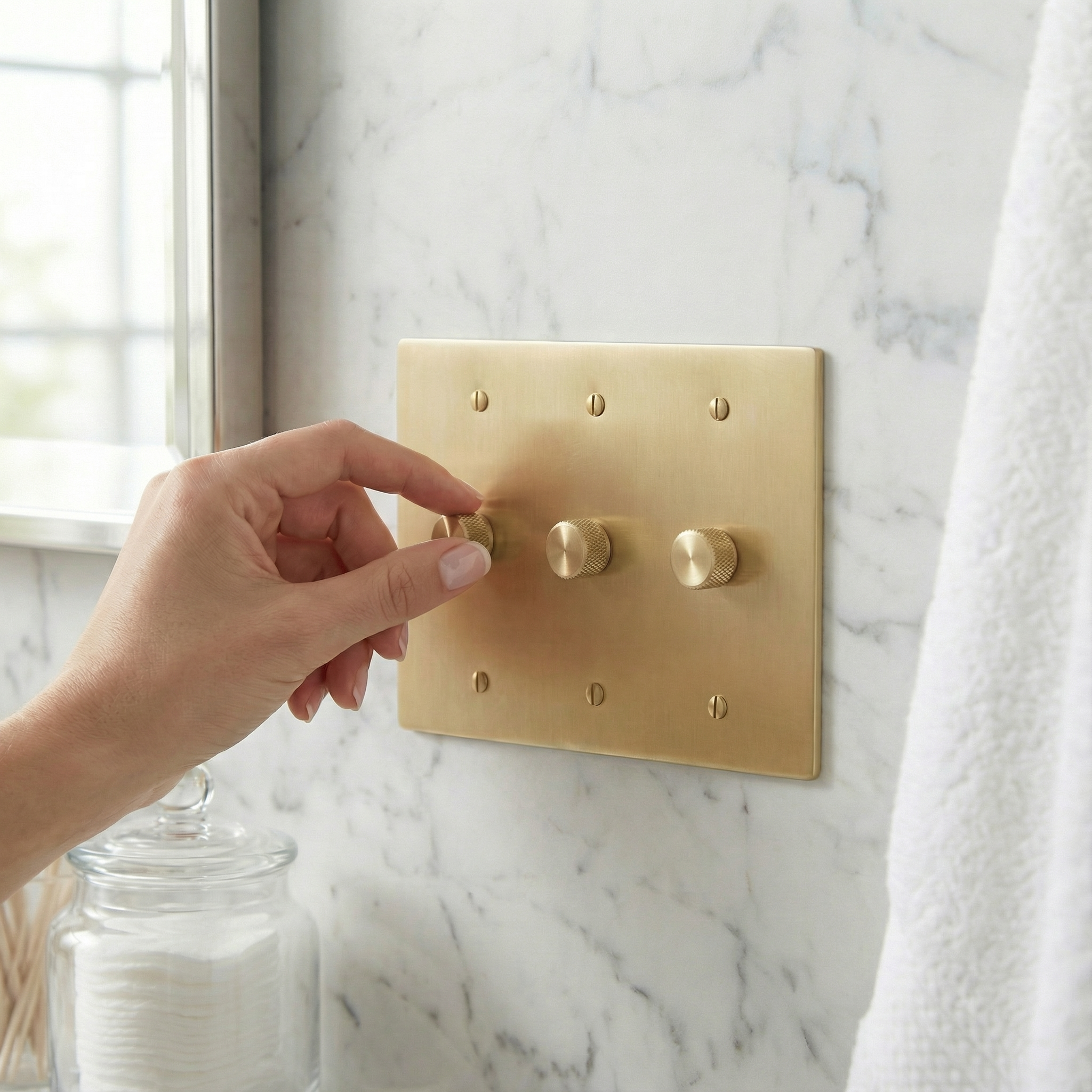 Hand adjusts a knurled knob on a satin brass 3 gang dimmer switch plate, blending luxury and timeless design on marble.