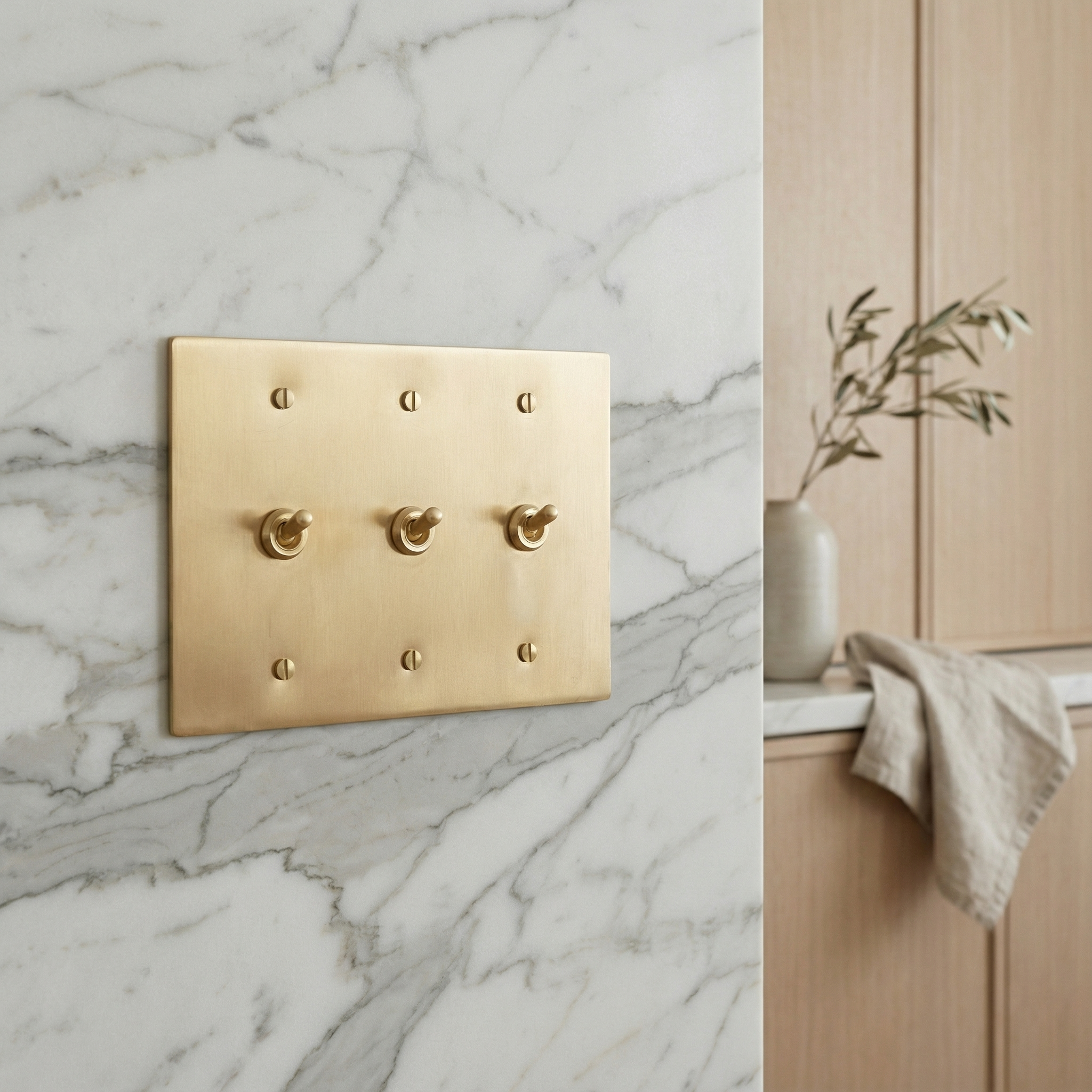 Satin brass 3 gang vintage toggle switch plate on a marble wall, evoking timeless elegance in a quiet luxury home setting.