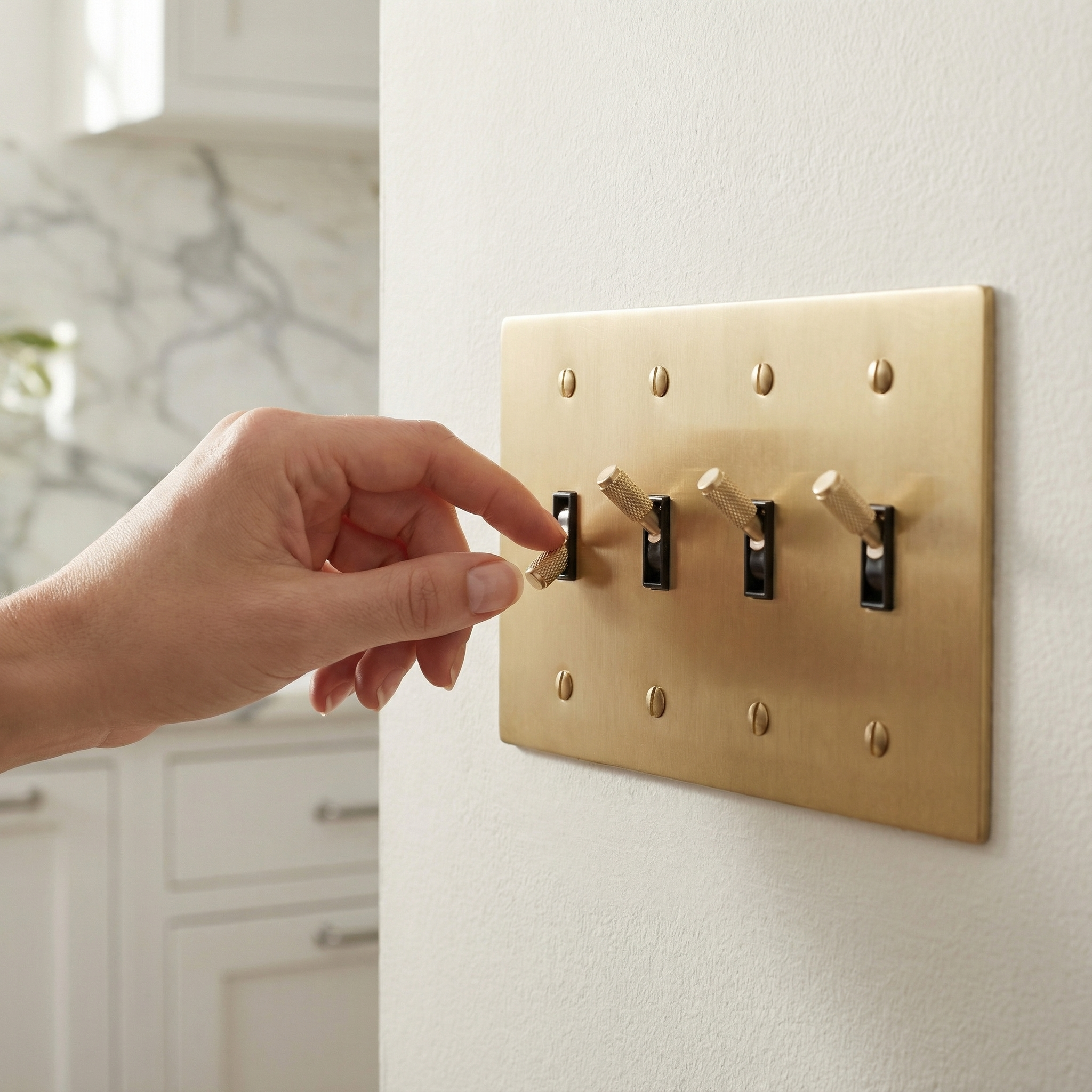 A hand flips a knurled toggle on a satin brass 4 gang toggle switch plate in a luxurious kitchen.