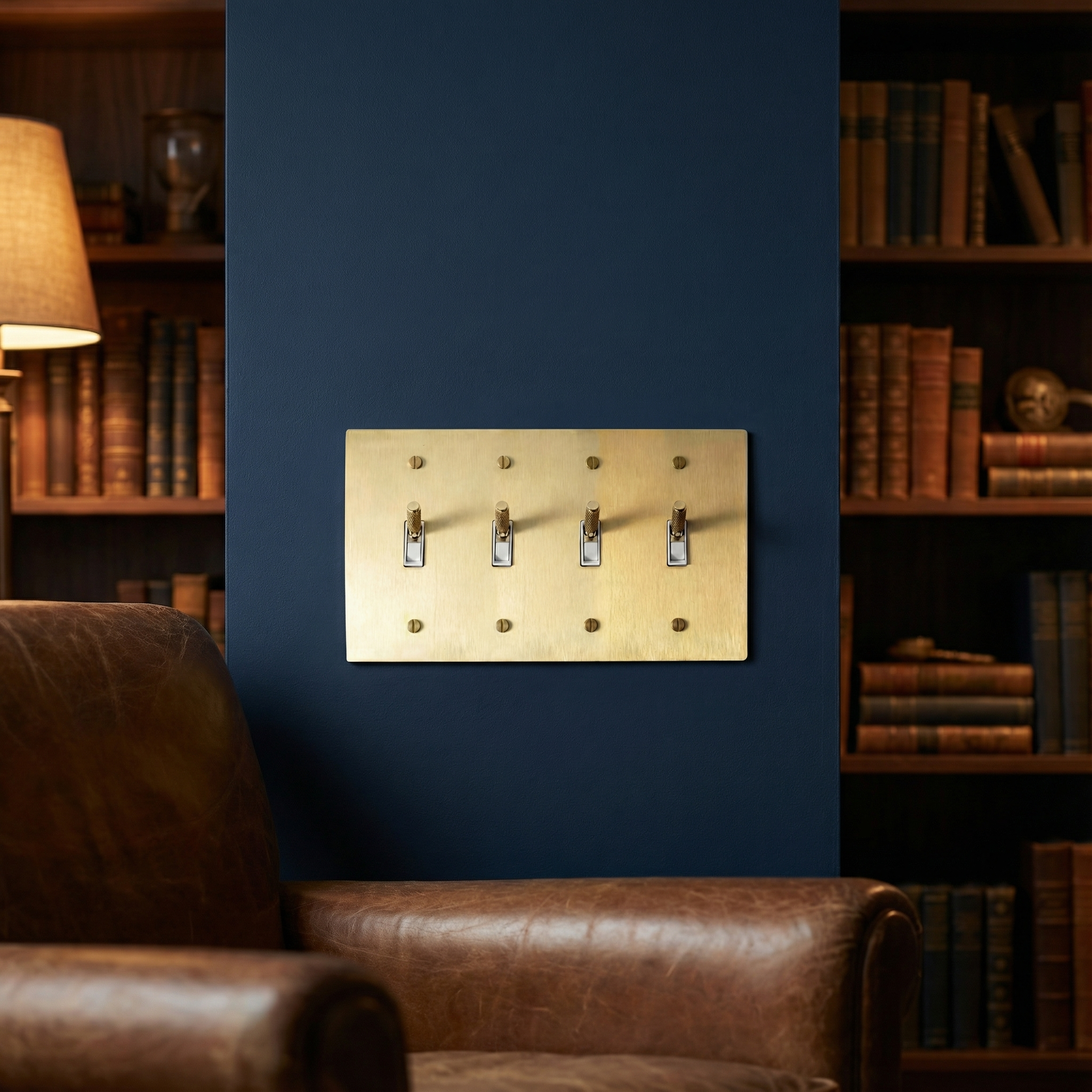 Satin brass 4 gang toggle switch plate on a deep blue wall, beside a leather armchair and bookshelves.