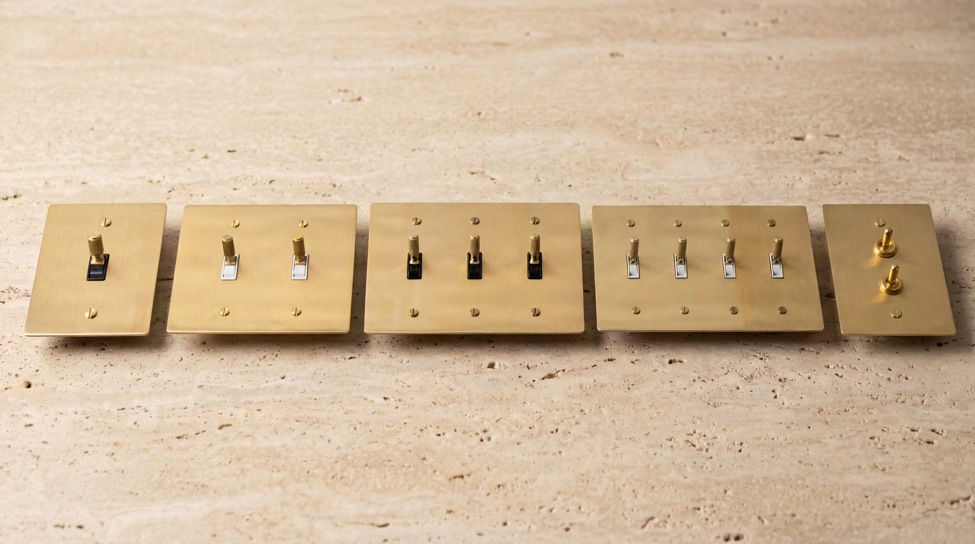 Satin Brass Toggle Switches: Five artisan-crafted solid brass plates, featuring knurled and smooth levers, on textured travertine.