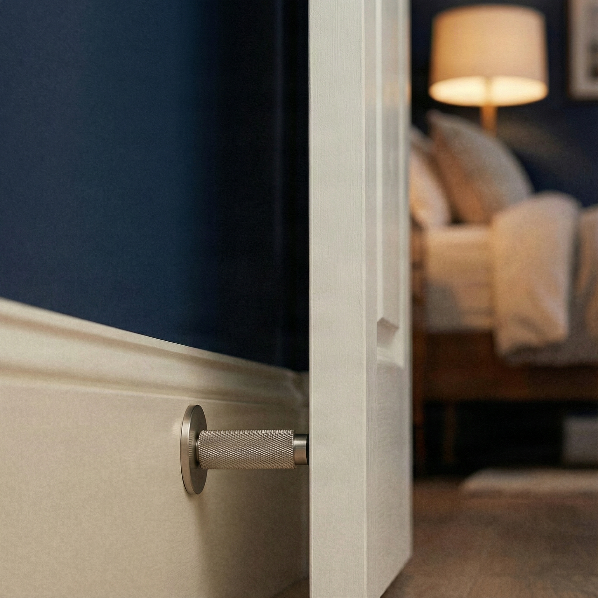 A modern satin nickel wall mount door stop secures an elegant white door against a deep navy wall, hinting at a serene bedroom beyond.
