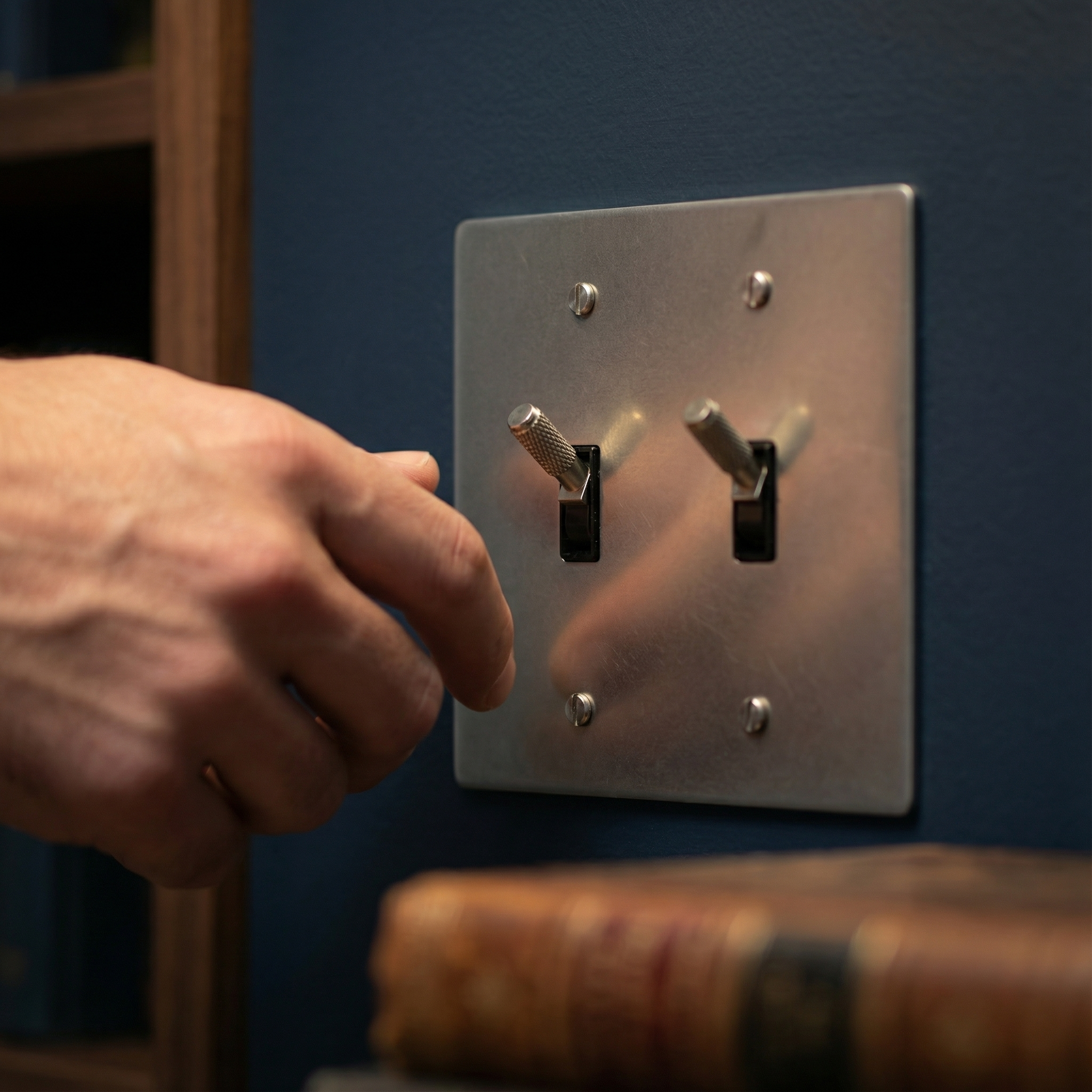 Hand reaches for a satin nickel 2 gang toggle switch plate on a dark wall, exuding quiet luxury.