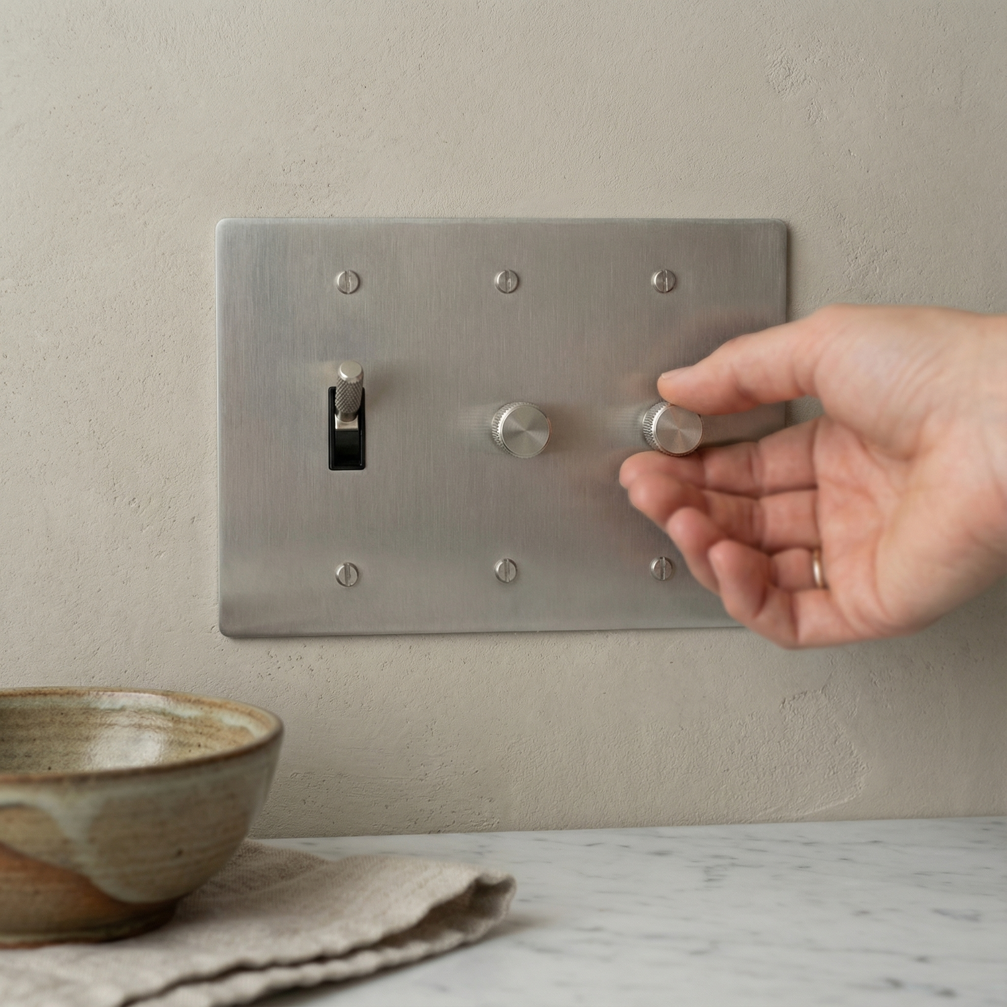 Hand adjusts a satin nickel 3 gang dimmer and toggle switch plate, exuding quiet luxury in a timeless setting.