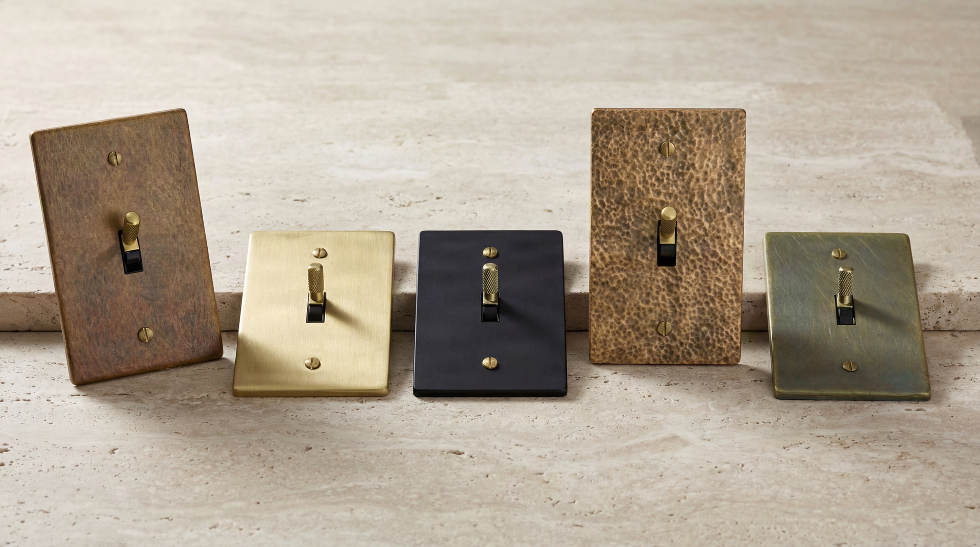 Five Metal Workz Toggle Switches in satin brass, aged, nickel, black, and antique finishes, with knurled and vintage levers, on stone.